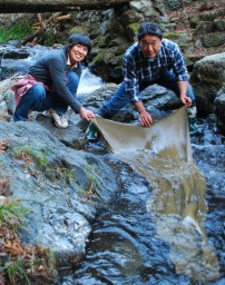 妻、ひろ子さん（左）と蚕のふんで染めた布を山田川に広げる松井さん
